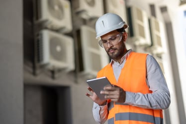 A professional wearing safety gear reviews data on a tablet in front of air conditioning units.