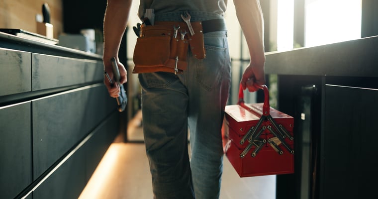 Rear view of a technician wearing a tool belt and carrying a red toolbox walking into a modern kitchen.